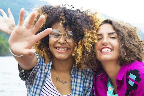 2 women waiving at camera and laughing while lakeside.